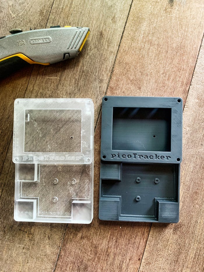 pexels photo 32764504 Display of transparent and black 3D printed PicoTracker cases next to a utility knife on a wooden surface.
